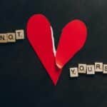Artistic image of a torn red heart with scrabble tiles spelling 'Not Yours' on a black background.
