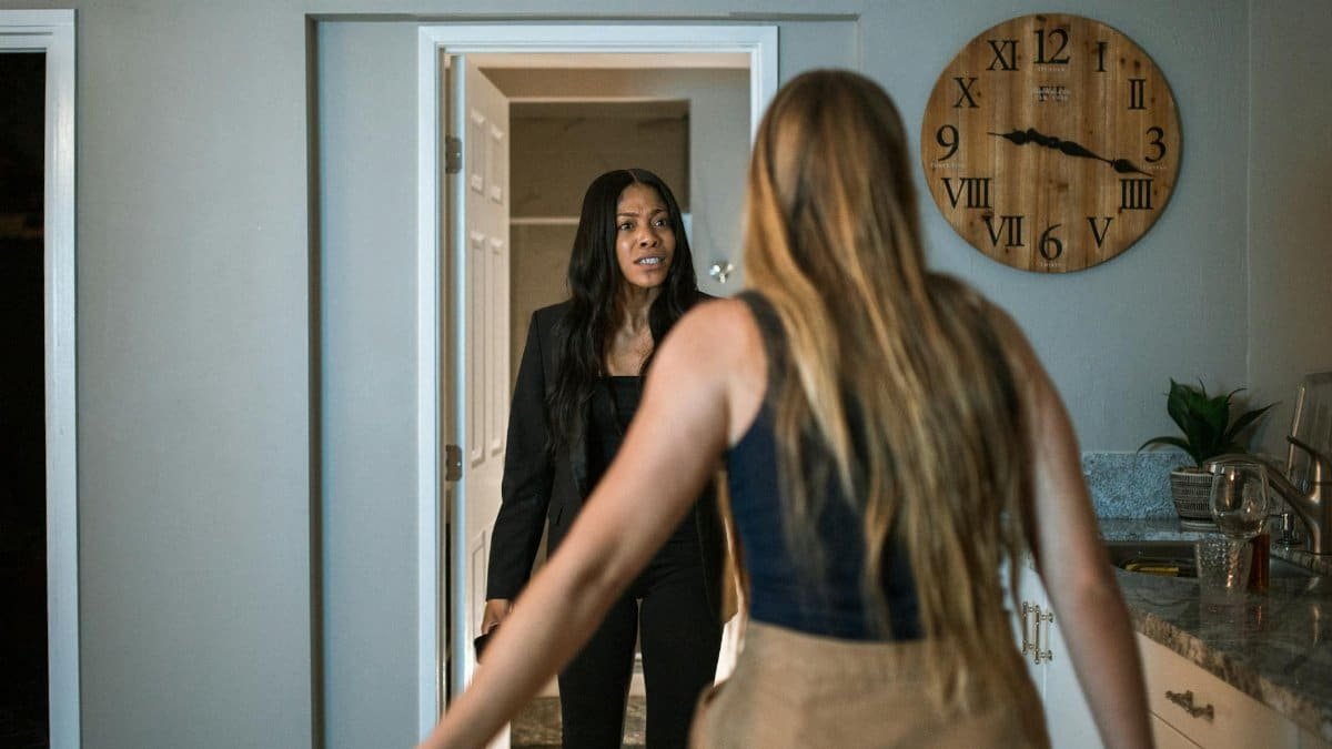Two women in a heated discussion inside a kitchen, next to a wall clock.