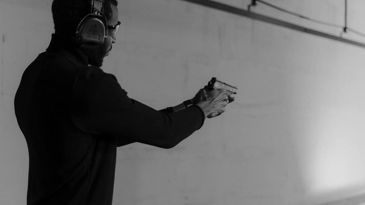 Black and white photo of a man aiming a pistol at an indoor firing range, wearing headphones.