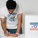 A man writes on a paper to cast his vote next to a ballot box in a studio setting.