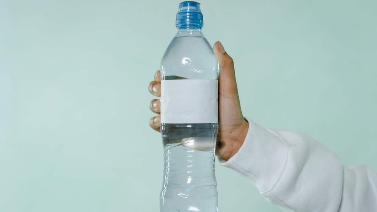 A hand holding a clear plastic water bottle with blank label on a soft green background.