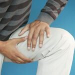 Close-up of a man's hands on his knee, showing pain or injury, on a blue background.