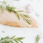 Healthy raw chicken breast garnished with rosemary on a marble surface.