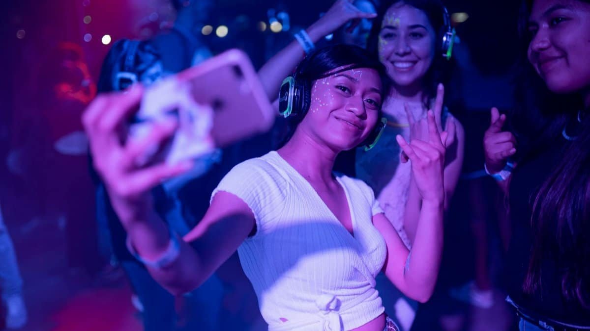 Young adults enjoying a silent disco party, capturing selfies amidst colorful lights.