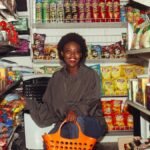 Full length of cheerful ethnic woman in casual clothes with shopping bag squatting among store shelves choosing food for buying