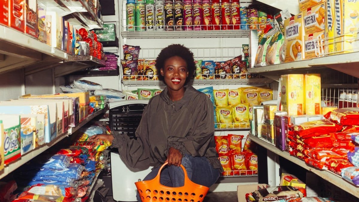 Full length of cheerful ethnic woman in casual clothes with shopping bag squatting among store shelves choosing food for buying