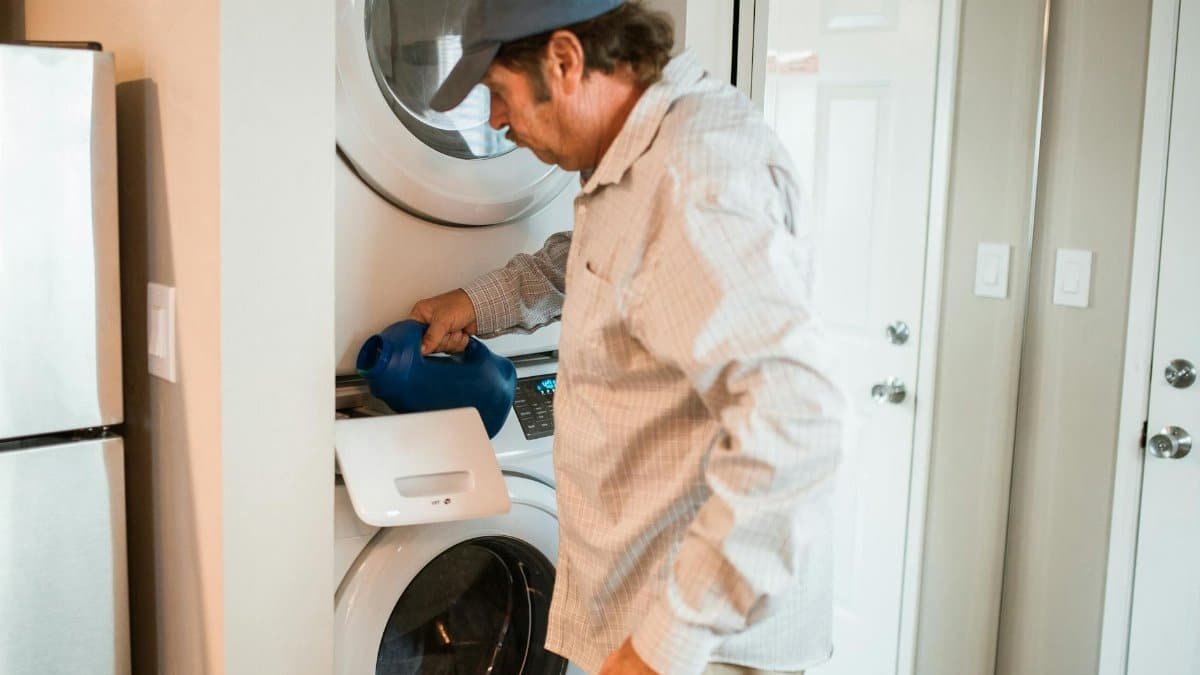 Man pouring liquid detergent into a stacked washer and dryer in a modern laundry room.