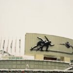 Sculpture of ice skaters at snowy winter sports venue, flags in background.