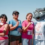 Four women celebrate with medals after a race alongside a sports mascot outdoors.
