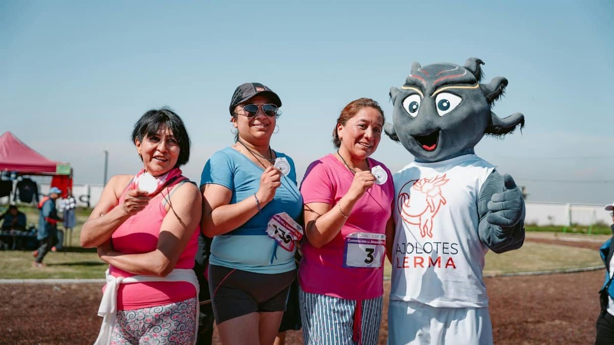 Four women celebrate with medals after a race alongside a sports mascot outdoors.