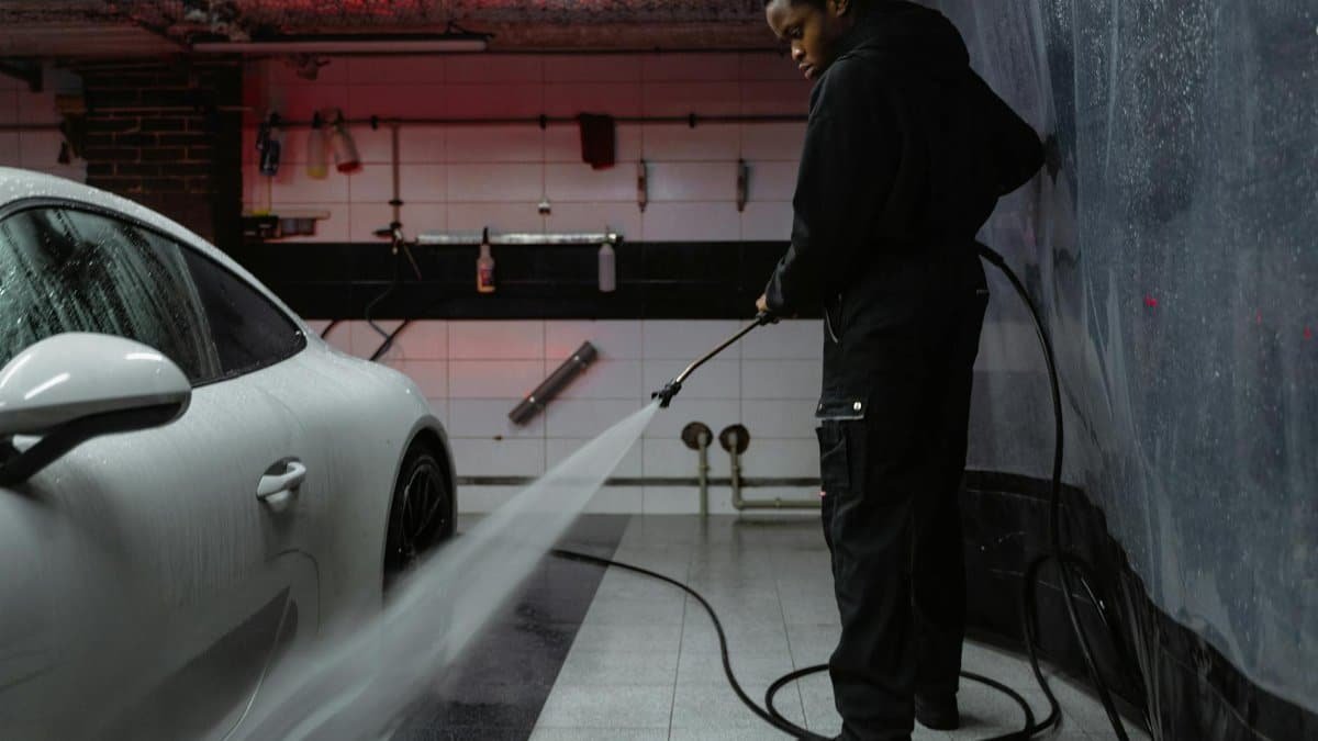 African American man cleaning a white car with a pressure hose in an indoor garage setting.
