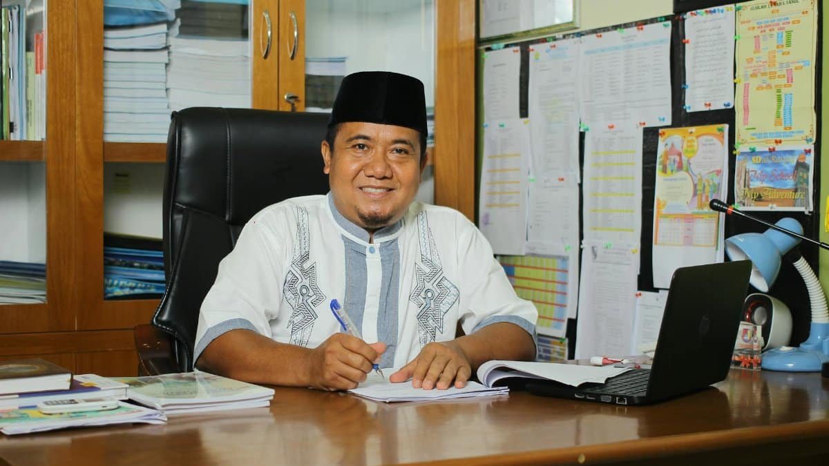 An Indonesian teacher in traditional attire, smiling at his desk with documents in an office setting.