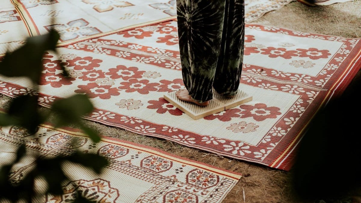 An individual stands barefoot on an acupressure mat surrounded by floral-patterned rugs, promoting relaxation.