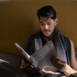 A man with dark hair reads a newspaper in bed during a quiet morning routine.