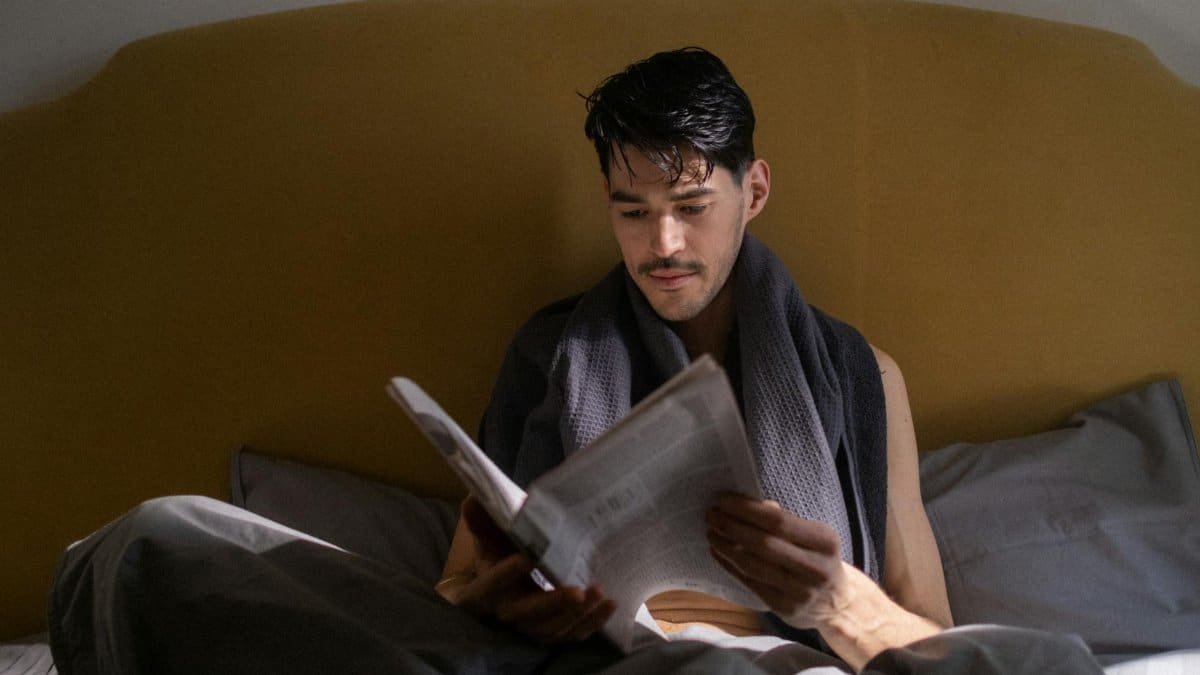 A man with dark hair reads a newspaper in bed during a quiet morning routine.
