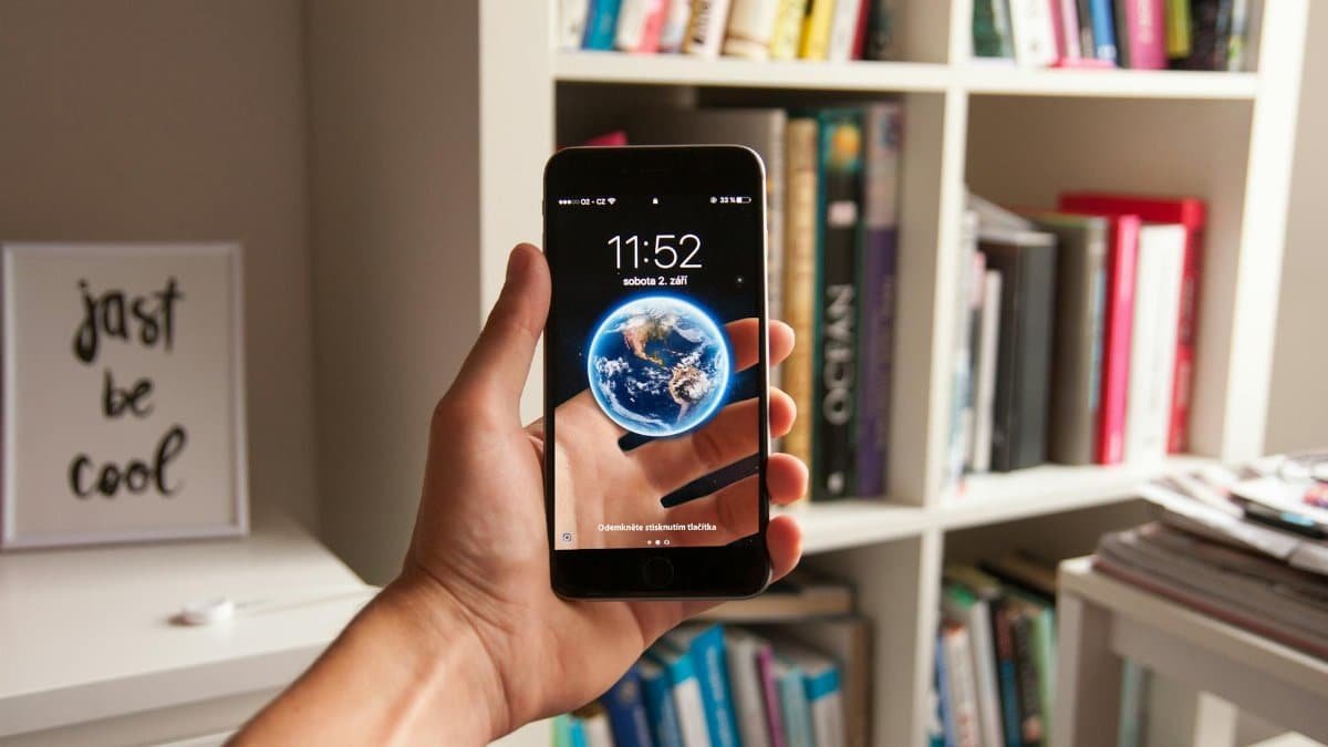 Hand holding smartphone with earth wallpaper, surrounded by books in a study room.