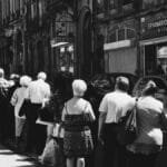 Monochrome street photo capturing a queue of people standing in front of old buildings in an urban setting.