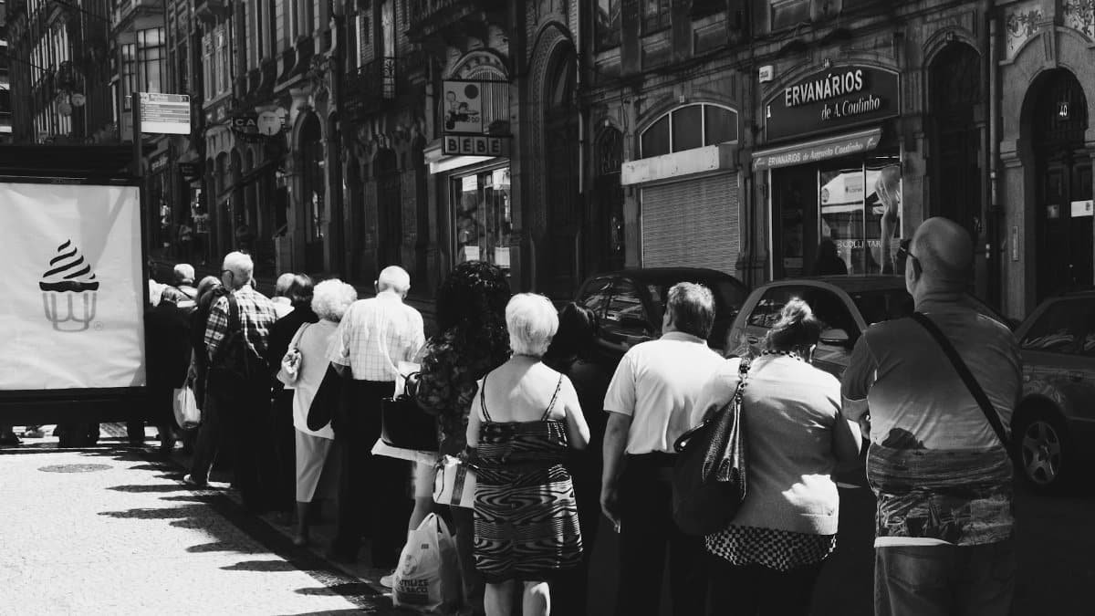 Monochrome street photo capturing a queue of people standing in front of old buildings in an urban setting.
