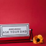 A whimsical Mother's Day sign with 'Mom's Mood: Ask Your Dad' text, set against a red backdrop with a flower.