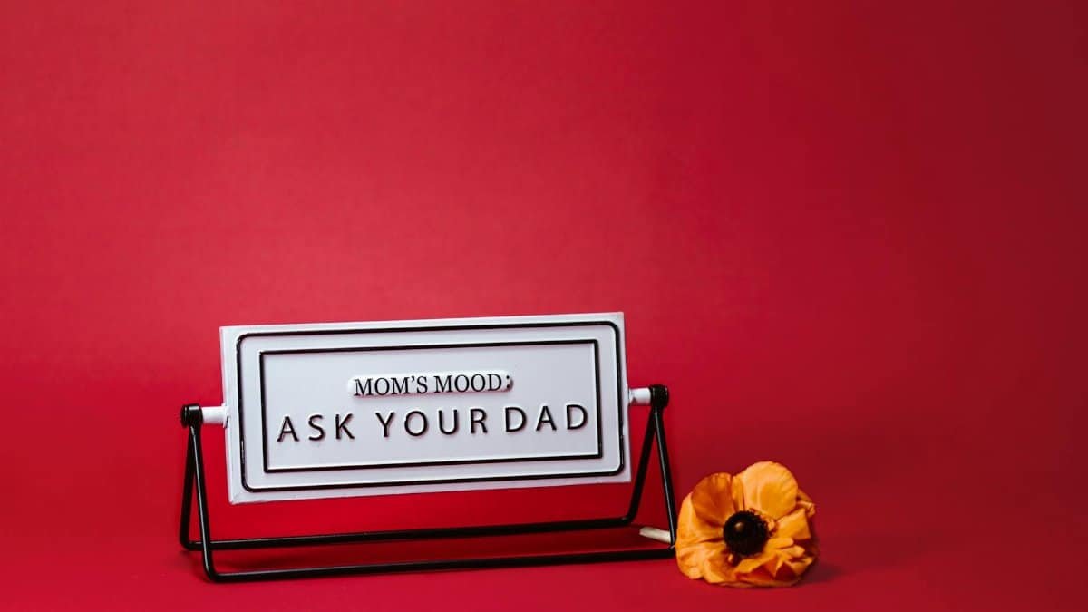 A whimsical Mother's Day sign with 'Mom's Mood: Ask Your Dad' text, set against a red backdrop with a flower.