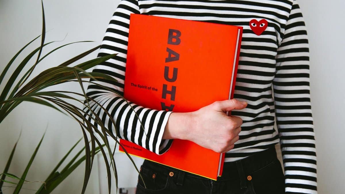 A woman with a striped top holds a Bauhaus book, embracing modern fashion.
