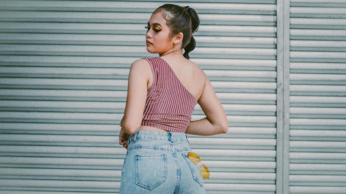Fashionable woman in denim and one-shoulder top posing outdoors by a roll-up door.
