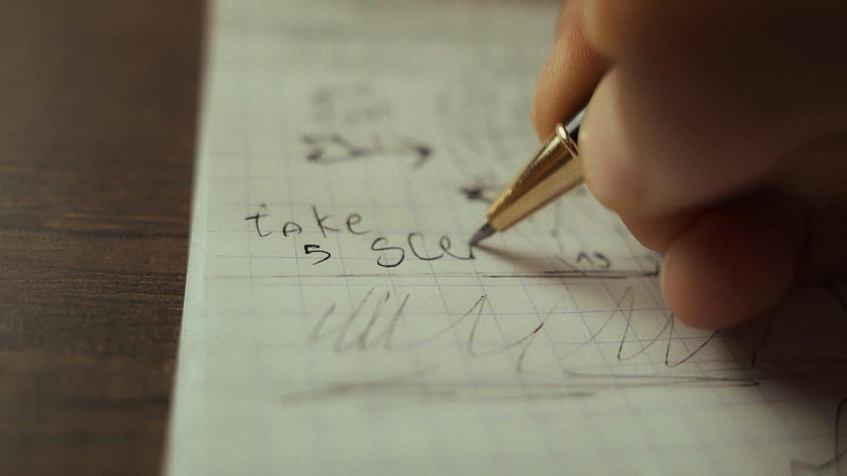 Close-up of a hand writing 'take 5 sec' on graph paper with a pen.