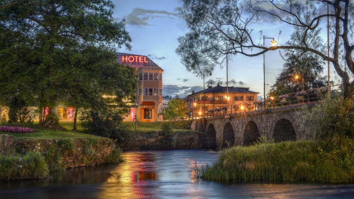 Charming twilight view of a hotel and stone bridge over a serene river with glowing lights and lush greenery.