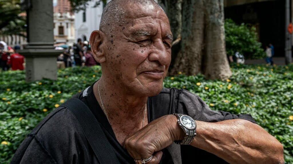 Elderly man with a watch sitting peacefully in a vibrant urban park near a large tree.