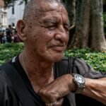 Elderly man with a watch sitting peacefully in a vibrant urban park near a large tree.