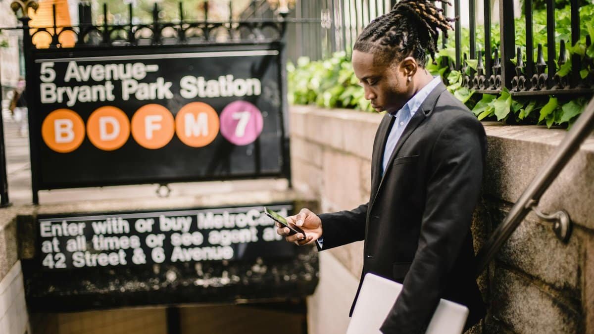 Side view of serious black man in office suit with dreadlocks and with laptop in hand texting on mobile while standing at subway entrance in Manhattan on sunny day