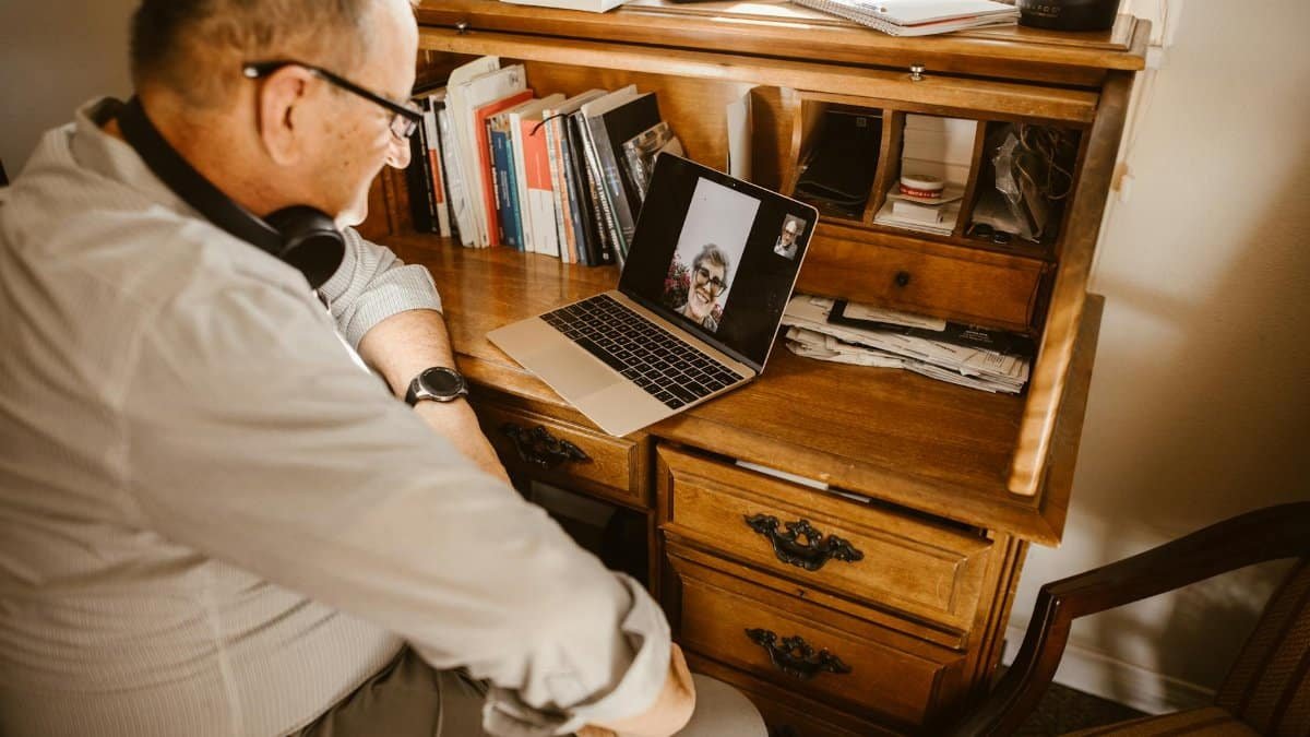 Senior man having a video call using a laptop at a wooden desk indoors.