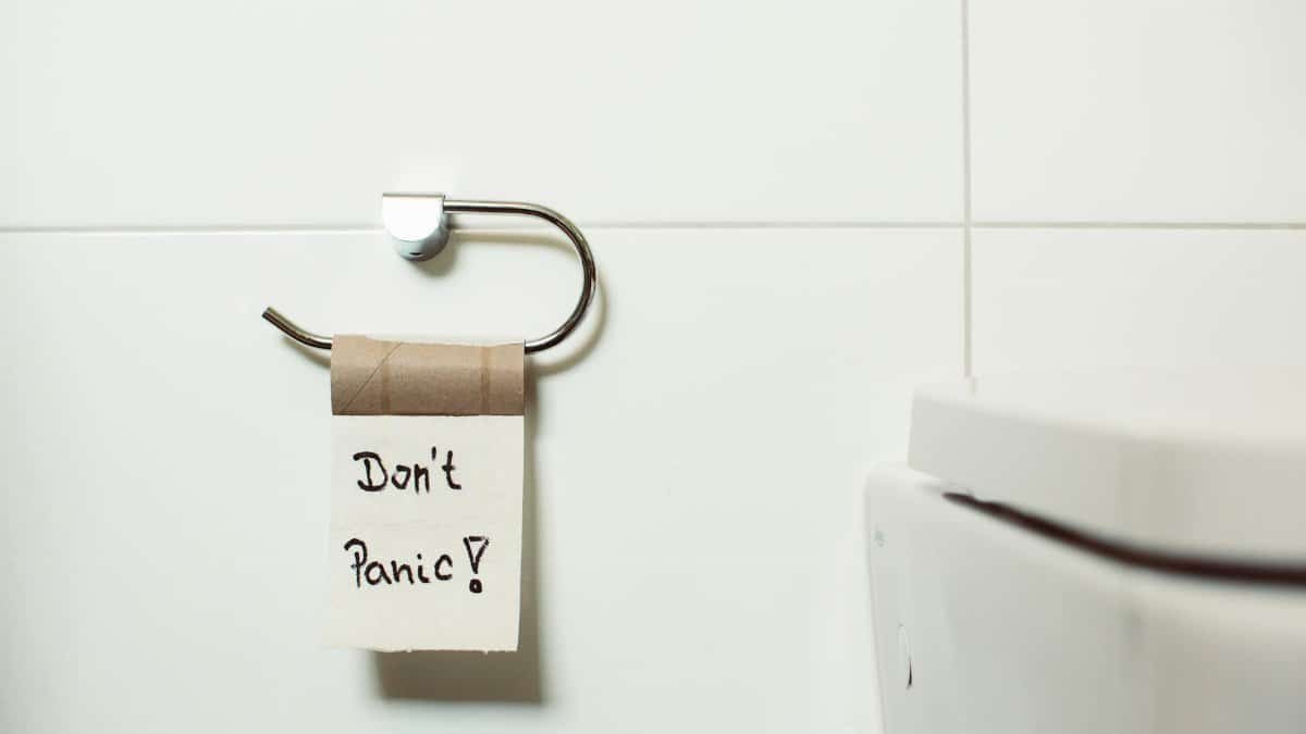 A toilet paper holder with an empty roll displaying a 'Don't Panic!' note, set in a modern bathroom.