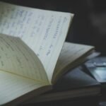 Open notebook with handwritten notes next to a mobile phone on a wooden desk in a dimly lit indoor setting.