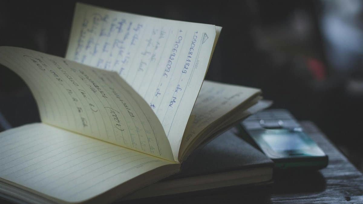 Open notebook with handwritten notes next to a mobile phone on a wooden desk in a dimly lit indoor setting.