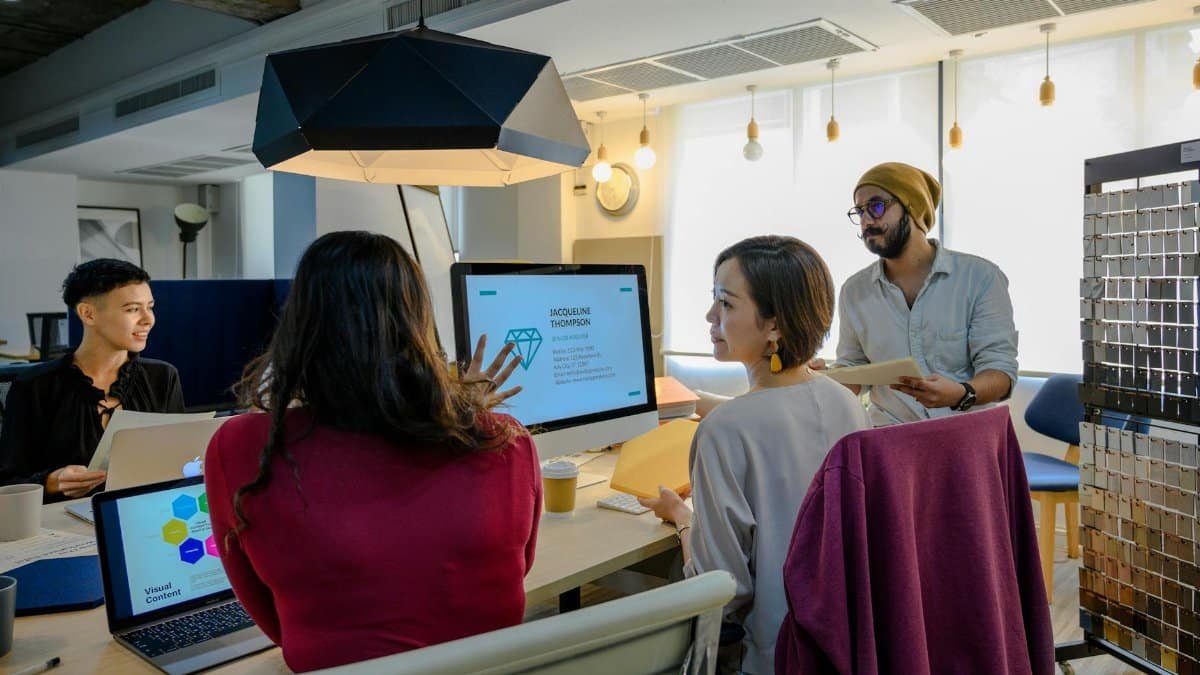 A diverse group of professionals working together in a contemporary office setting using laptops and a monitor.