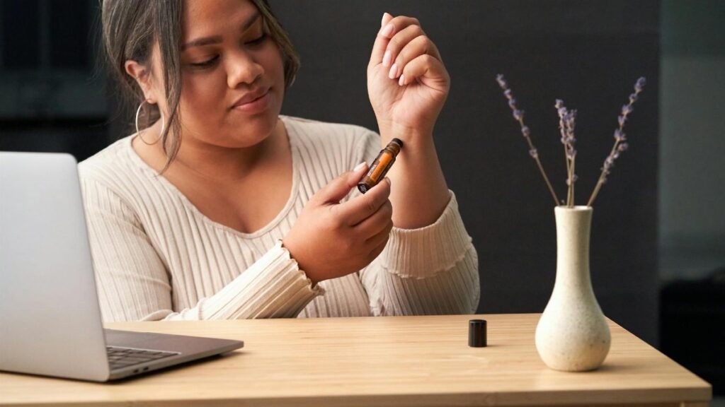 A woman uses a therapeutic essential oil roll-on at a work desk featuring a laptop and vase with flowers.