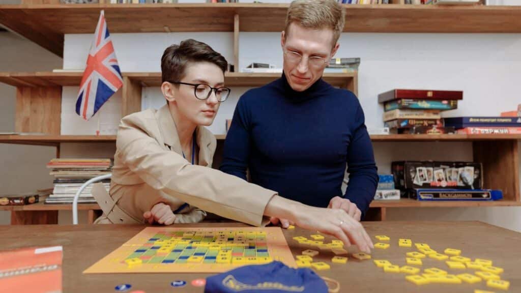A man and woman play a board game together in a cozy library setting with books and a flag.