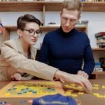 A man and woman play a board game together in a cozy library setting with books and a flag.