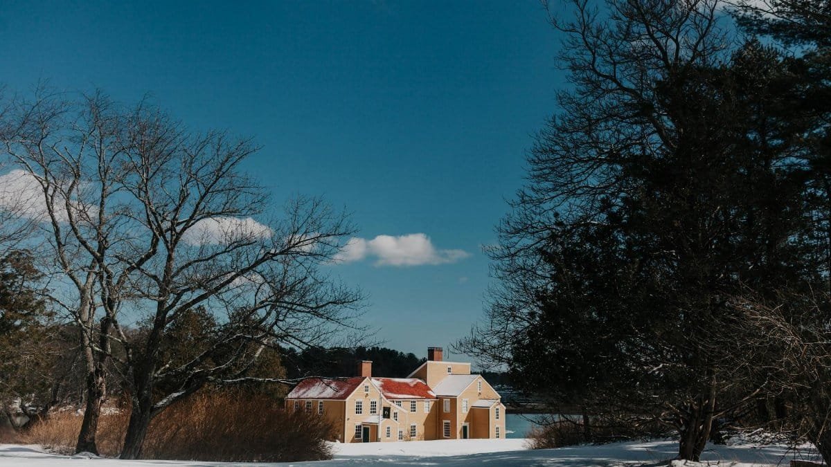 Winter scene with a quaint yellow house surrounded by snow and bare trees in New Hampshire.
