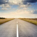 A long straight road stretches through fields under a dramatic cloudy sky.