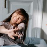Smiling Asian woman stretching in bed, enjoying soft morning light indoors.