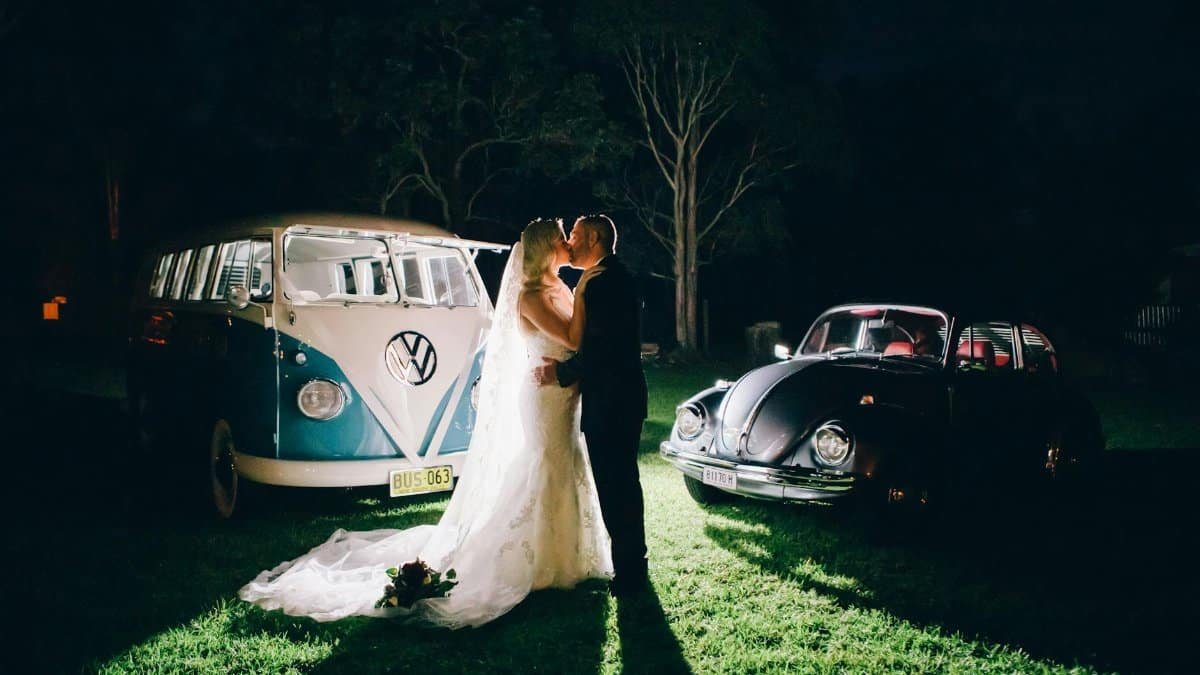 A bride and groom share a kiss beside vintage VW cars during a rustic nighttime wedding in Australia.