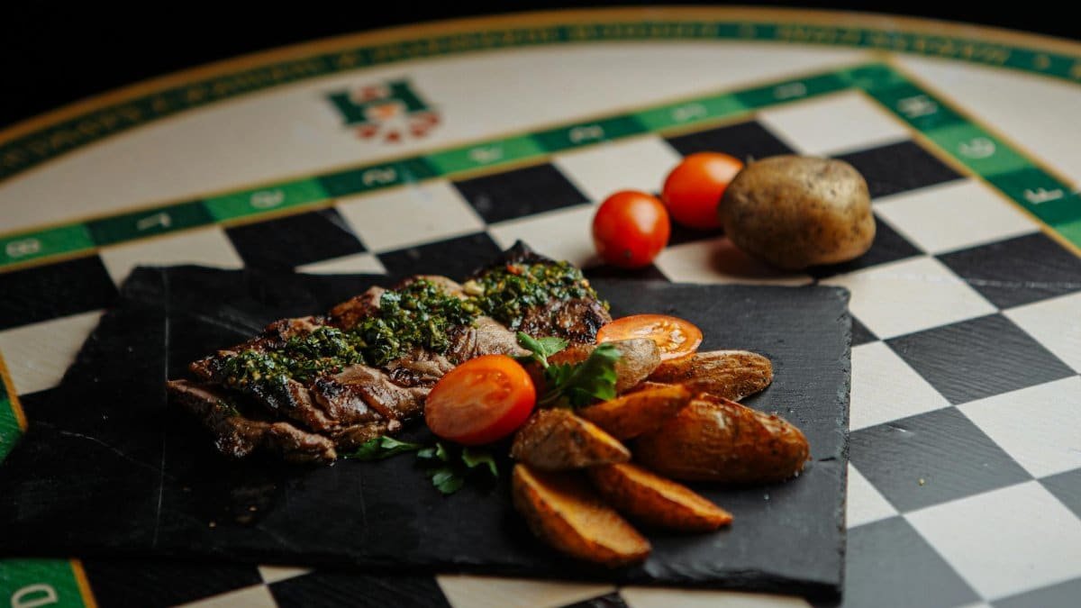 Sizzling grilled steak with chimichurri, served with roasted potatoes and fresh tomatoes on a slate board.