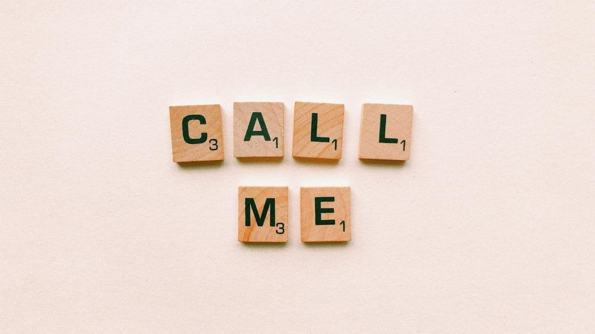 Wooden letter tiles spelling 'Call Me' on a pastel pink background, invoking communication and connection.