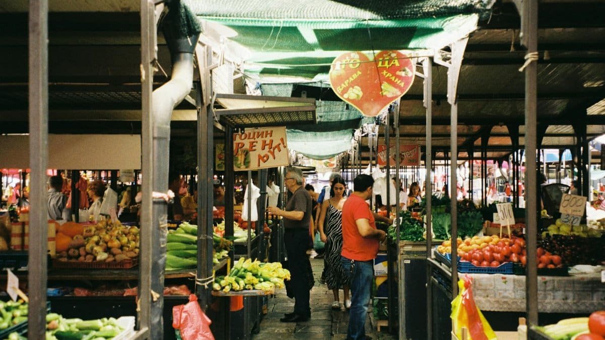 A bustling outdoor farmer's market with fresh vegetables and people shopping on a sunny day.