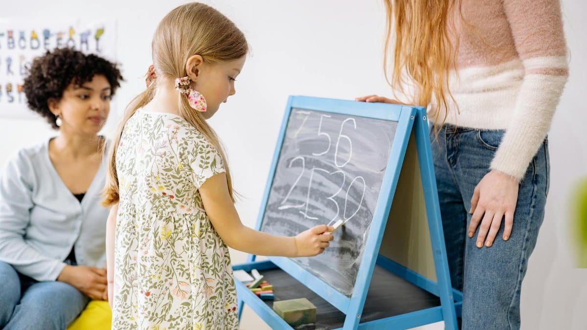 A young girl writes numbers on a chalkboard in a classroom, supervised by adults.