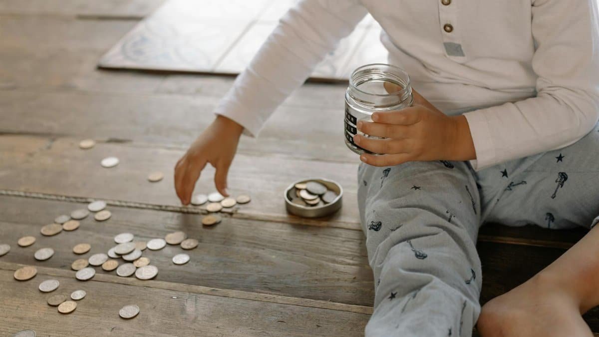 A young child collects coins into a jar on a wooden floor, symbolizing savings.