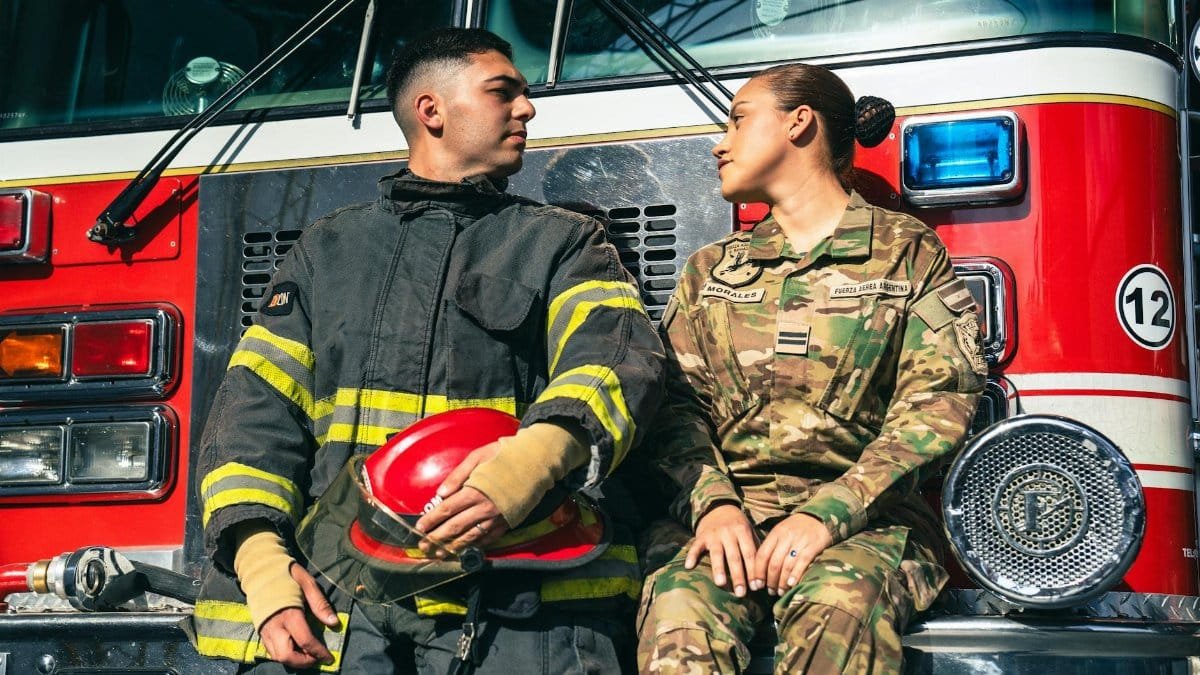 A firefighter and a soldier sit in front of a fire truck, sharing a candid moment.