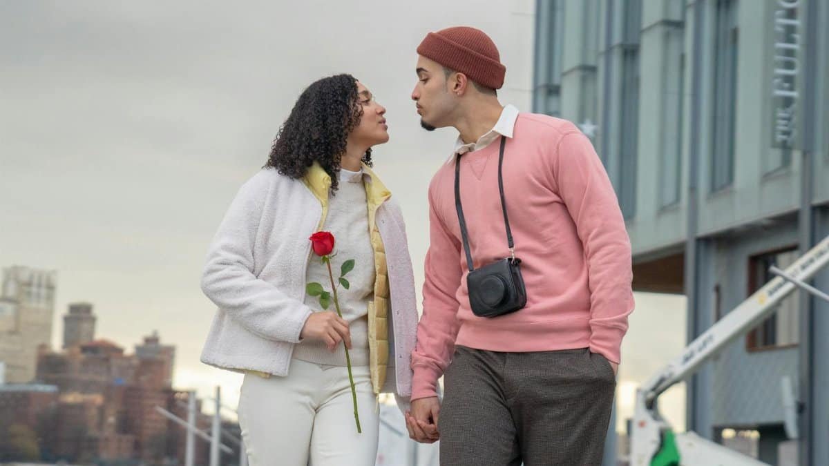Loving Hispanic girlfriend with flower and boyfriend with photo camera holding hands and looking at each other while strolling on street